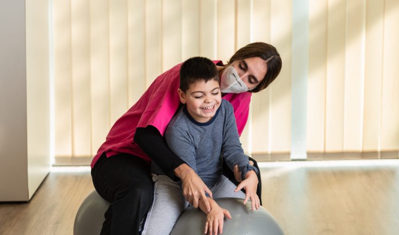 child with multiple intellectual and physical disabilities and physiotherapist woman do balance exercises on a Peanut Gym Ball with joy. she wears coronavirus pandemic protection mask