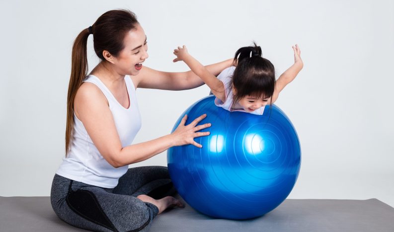 Cheerful asian young mother and little daughter doing gymnastics exercise with fitness ball on white background