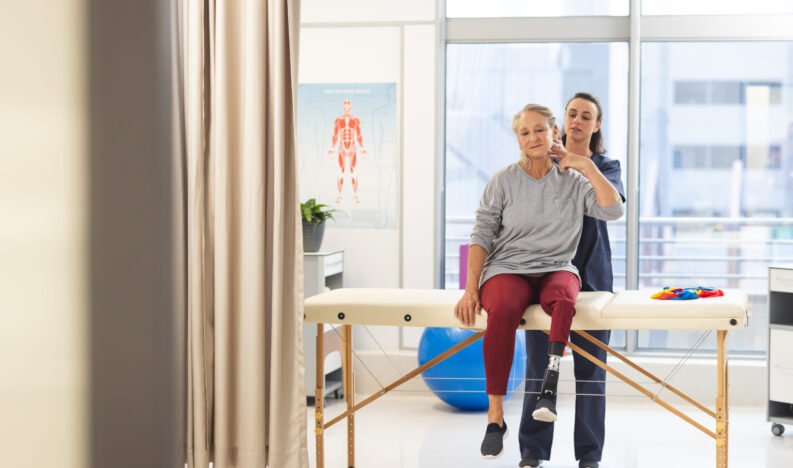 Caucasian female physiotherapist and senior woman with artificial leg massaging back at hospital. Hospital, disability, physiotherapy, work, medicine and healthcare, unaltered.