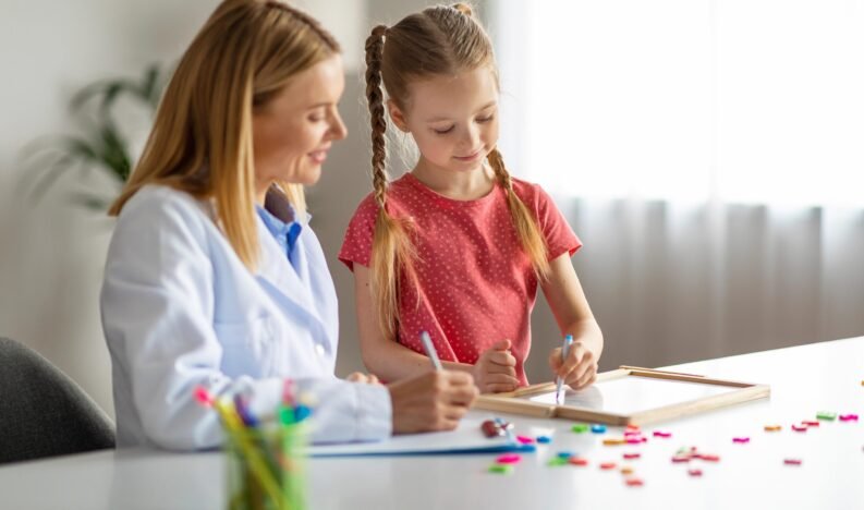Cute Little Girl Having Therapy Meeting With Professional Child Psychologist Woman, Smiling Female Kid Sitting At Table And Writing On Drawing Board, Learning Letters, Treating Dyslexia, Copy Space