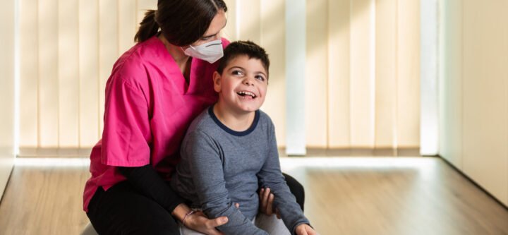 child with multiple intellectual and physical disabilities and physiotherapist woman do balance exercises on a Peanut Gym Ball with joy. she wears coronavirus pandemic protection mask