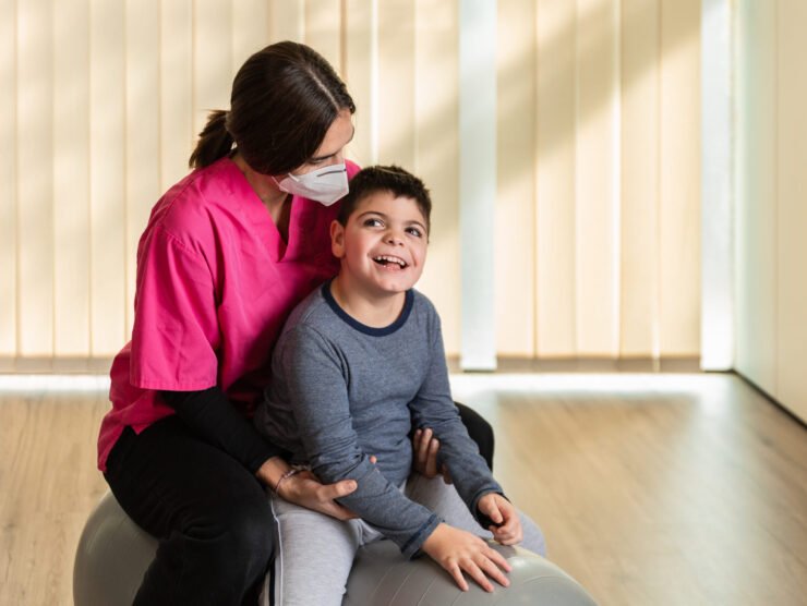 child with multiple intellectual and physical disabilities and physiotherapist woman do balance exercises on a Peanut Gym Ball with joy. she wears coronavirus pandemic protection mask