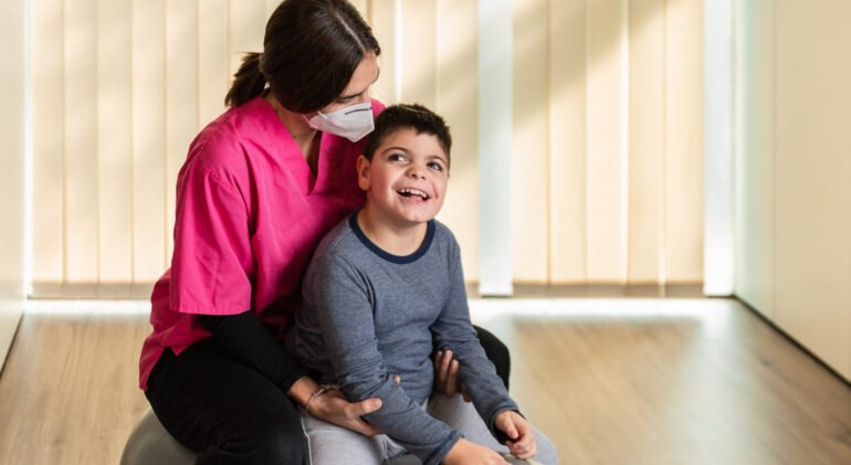 child with multiple intellectual and physical disabilities and physiotherapist woman do balance exercises on a Peanut Gym Ball with joy. she wears coronavirus pandemic protection mask