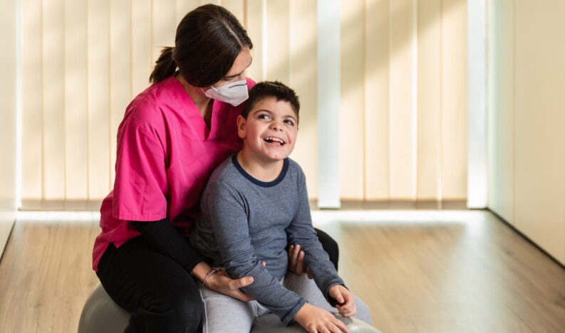 child with multiple intellectual and physical disabilities and physiotherapist woman do balance exercises on a Peanut Gym Ball with joy. she wears coronavirus pandemic protection mask